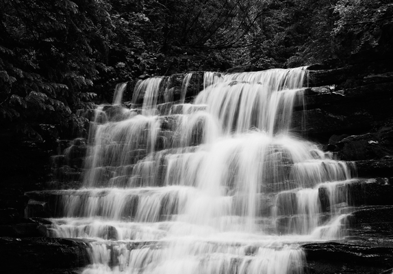 A black and white image of a cascading waterfall flowing over a series of rock ledges. The water appears silky and smooth, contrasting with the dark, textured rocks. Surrounding the waterfall are dense trees and foliage, adding depth and framing the scene in a natural setting.