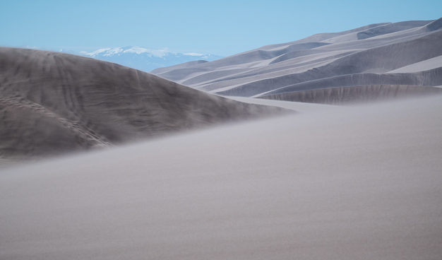 Sweeping sand dunes stretch across the landscape under a clear blue sky. The dunes are smooth and undulating, with strong winds lifting fine sand into the air, creating a hazy effect near the ground. In the distance, snow-capped mountains rise, contrasting with the arid desert foreground.