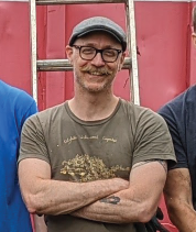 Photo of me, a white, bald man in my 40s. I'm mustachioed, wearing glasses and a gray, flat cap (english style). I have my arms crossed and I'm smiling. I'm standing in front of a ladder leading into a dumpster.