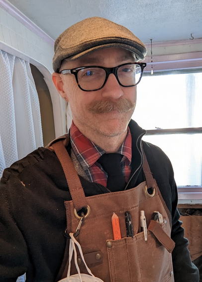 Photo of me, a white, bald man in my 40s. I have a thick mustache and I'm wearing a gray flat english cap and glasses. I have on a checkered shirt and black tie as well as a black sweater. Over top of that I have a tan work apron with tools in the pockets. Over my shoulder you can see a room under construction.