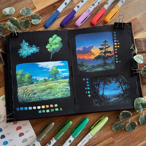 A photo of a black paper sketchbook placed on a wooden table, surrounded by swatches, acrylic markers, and leaves for decoration. On the left page you can see some foliage and a simple tree, and underneath a meadow in green/brown tones under a bright blue sky with a tree line in between. On the top half of the right page you can see an evening sky, part blue part orange, and a winding path in drab teal tones. The silhouette of a pine tree is visible on the right side. On the bottom half of the right page you can see a nighttime sky and some grass through the black shapes of tree trunks, foliage, and shrubs. Colour swatches are painted next to each scenery.