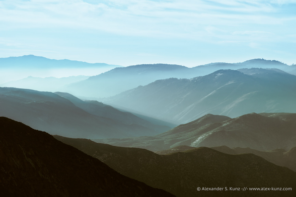 A landscape photo showing a series of interesection mountain ridges receding into afternoon haze. The sky above the mountains has faint, hazy clouds in it. The color palette is a mix of dark brown tones in the foreground to light blue in the distance.