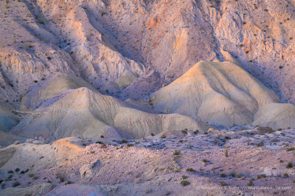 An intimate landscape photo showing a barren desert landscape, indirectly illuminated by the evening sky after sunset. The prominent feature are two mud hills, embedded in sparsely vegetated rocky hillsides. The color palette includes pale beige, orange and purple as the landscape takes on the colors of the clear evening sky.