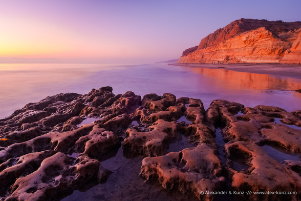 A photo showing a seascape after sunset. Looking north with the camera, indirect light from the evening sky in the west (left in the frame)  illuminates a cliff in warm orange tones. In the immediate foreground, a heavily eroded rock has water in shallow depressions. The water of the ocean is rendered smooth with a long exposure time.