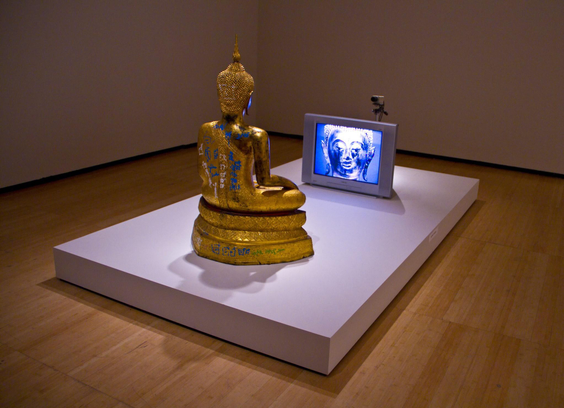 Color photo of a Nam June Paik closed circuit Buddha. Looks like a golden Thai Buddha sitting on a white platform facing a television with a camera on top that is filming him as he sits in mediation. 