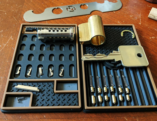 A photo of a fully picked and gutted challenge lock. The lock has been laid out on two black Sparrows pinning mats. The core, sidebar, sliders, gin-bottle driver and key pins, the housing, springs, and key are neatly arranged. There is a Sparrows gutting tool (that looks like a tiny Klingon weapon) placed above the mats.