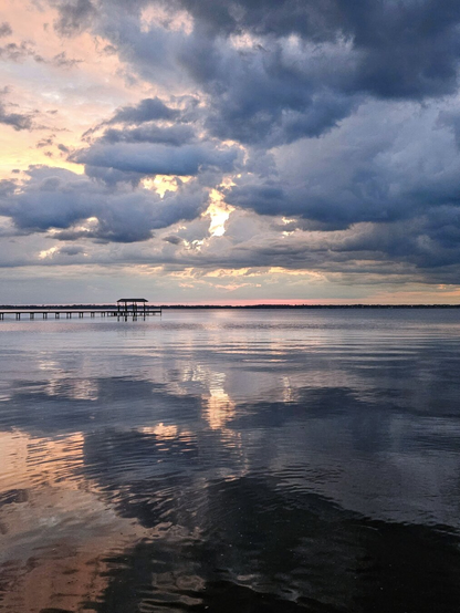 Pastel colors overtake the big Florida sky as storm clouds move in over a vast river just as the sun is lowering near the horizon.  The sun's intensity manages to cast shades of pink, yellow, and purple into the spaces between blue-grey cloud formations. The incredible colors and shapes are all reflected back by the calm, almost mirror like river's surface.
