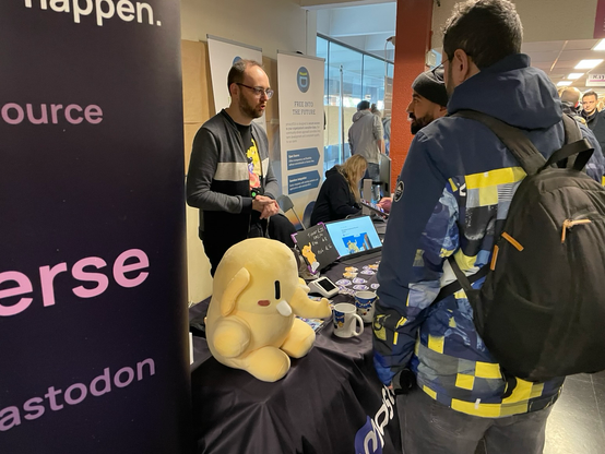 Mastodon stand with with the stuffed toy in the front, other merch in the back. Renaud is behind the stand. 