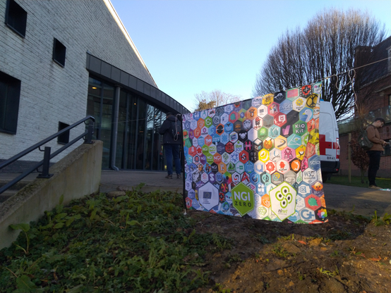 Flag hanging from a line with hex shaped logos of FOSS projects funded by NLnet. Left are the stairs that lead up to the K-building Level 2.