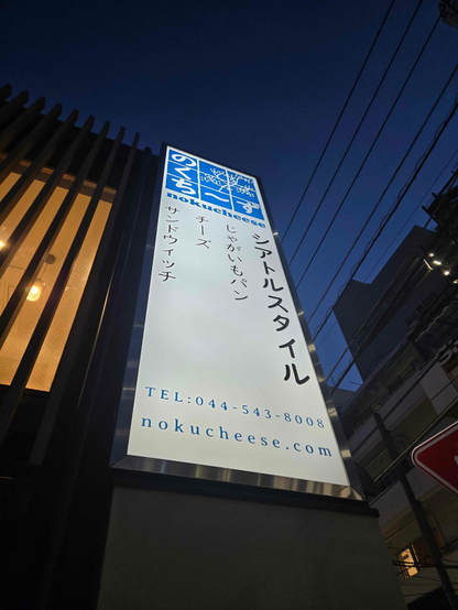 Vertical signage for a bakery/cafe named Nokucheese in Japan. The sign is composed of a logo at the top (described in more detail in the following attachments), some of the main items for sale (Seattle-style potato bread, cheese, and sandwiches), and the cafe's phone number and website address.