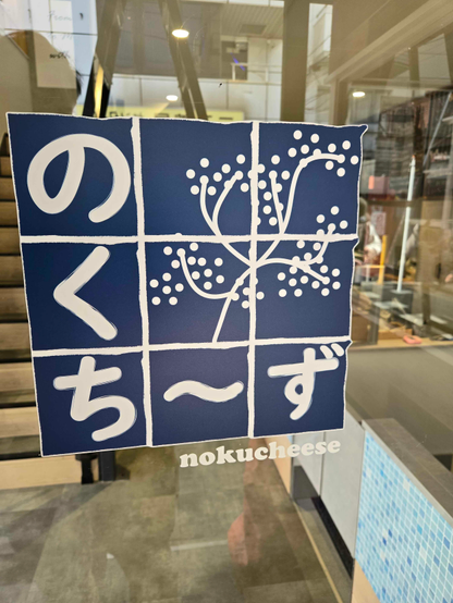 Photo from the interior of the cafe, closeup on the Nokucheese logo. The logo is a a series of indigo blocks with rough edges in a 3x3 grid. In a white color on top of the indigo blocks, the hiragana characters の・く・ち・～・ず are placed in an 'L' shape. In the remaining four blocks, a stylized herb with branches and small bulbs, also in white.