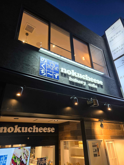 The outside of the cafe with the 3x3 indigo logo and the text, in English, "nokucheese", "bakery cafe" in cutout white letters. The storefront lighting creates a soft shadow of the lettering.