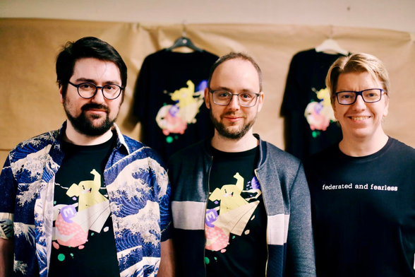 A photo showing the Mastodon team: Eugen Rochko with Renaud Chaput and Andy Piper. Behind them you can see Mastodon t-shirts for sale 
