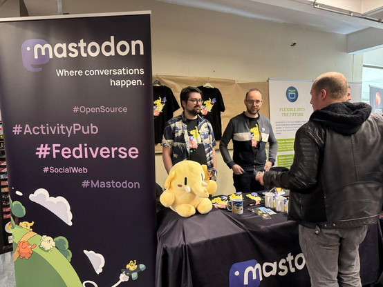 Photography of the Mastodon table at FOSDEM, featuring Renaud, Eugen and the Mastodon plushie throning on the table
