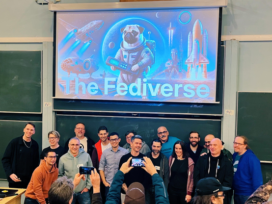 A photo of a large group of people (basically, Fediverse and ActivityPub royalty) standing in front of a slide from John O’Nolan talk about Ghost that shows their “ActivityPug” (an AI-generated pug in a space suit) with the word The Fediverse under it