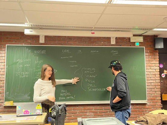 The image depicts two individuals,  engaged in a discussion in a classroom at Université Libre de Bruxelles. In the foreground, there's a blackboard filled with technical notations, and in the background, there's a brick wall.