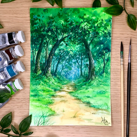 A photo of a painting placed on a wooden table, surrounded by paint tubes and brushes, as well as some leaves for decoration. The painting features a grass-rimmed packed dirt pathway leading into bright woods. The predominant colour is green; the light in the forest is pale bluish.