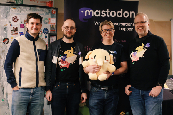 A portrait of the Mastodon team standing in front of their stand at FOSDEM. From left: Felix, Renaud, Andy (holding the Mastodon plushie) and David. Behind them there is a banner that reads “Mastodon - where conversations happen”