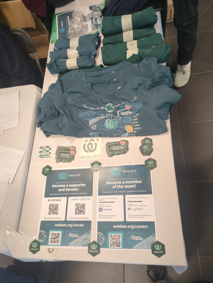 Portrait oriented photograph of the weblate stand display at FOSDEM 2025. There are 2 posters at the bottom of the table, t-shirts at its top and many stickers witch 4 different designes in between.