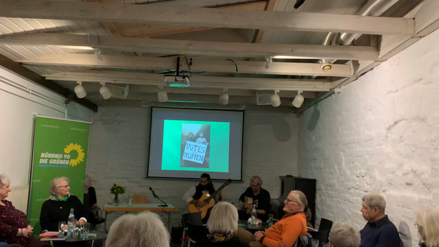 Coskun Wuppinger Duo playing jazz on guitars. The audience is listening to their music. In the background is a banner for Bündnis 90/Die Grünen. A black white photograph “woman voters” is projected onto a wall