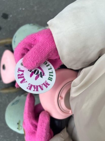 a photo of my daughter's hands (she is wearing fuchsia gloves) holding the sticker "make art not billionaires" on our way to pre-school