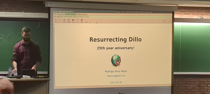 The image shows a man giving a presentation on a screen. The screen displays the title "Resurrecting Dillo", the subtitle "25th Anniversary!".
The man is standing in front of the screen, with a laptop on a table in front of him. Behind him, there is a green chalkboard.