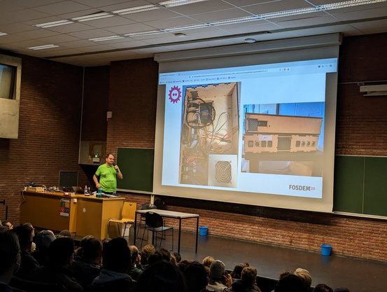 Basti on stage, with a slide showing FOSDEM's video boxes from the inside, the side, and the front.