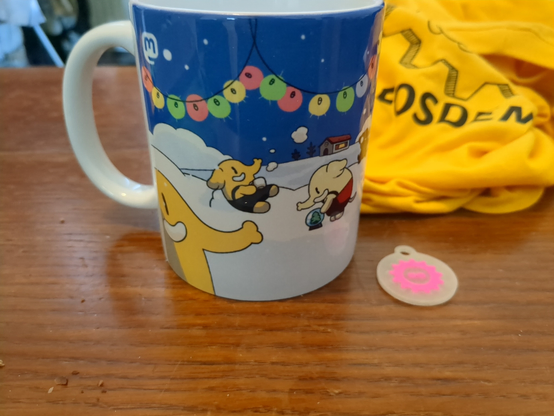 sur une table basse, un mug mastodon avec des mastodon qui jouent dans la neige, un goodies fosdem (exclusif je pense ^^ ) et mon pull qui traîne derrière