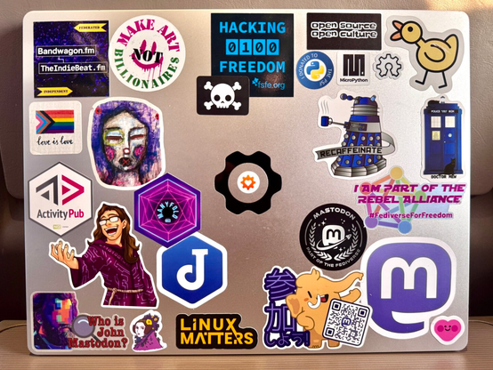 The lid of a silvery 14 inch Framework laptop covered in stickers, including the logos of many #Fediverse-related technologies and platforms. There is a cluster of Mastodon stickers in the bottom right corner of the case.