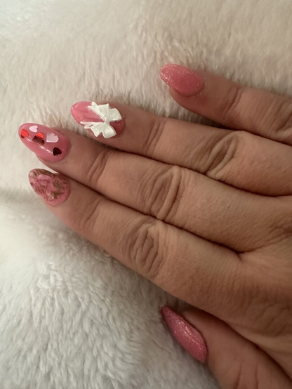 A picture of my manicure that I got for Valentine’s Day. The nails are pink. The thumb and pinky have glitter. The pointer has a rose decal. The middle finger has confetti hearts. The ring finger has a white 3D bow.