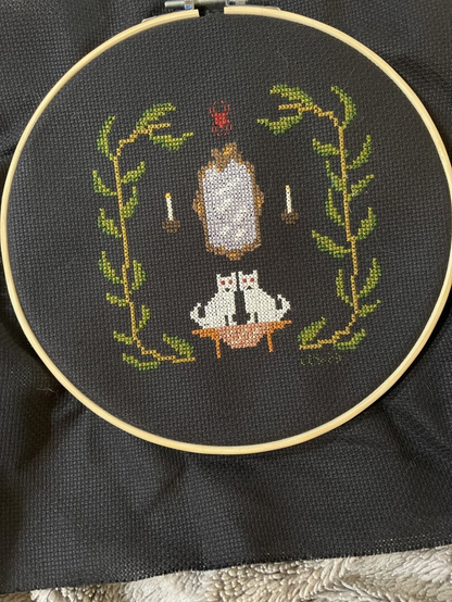 Completed cross-stitch on black Aida of two white kitties sitting underneath a mirror with vines on both sides, making a frame. From Creepy Cross-stitch