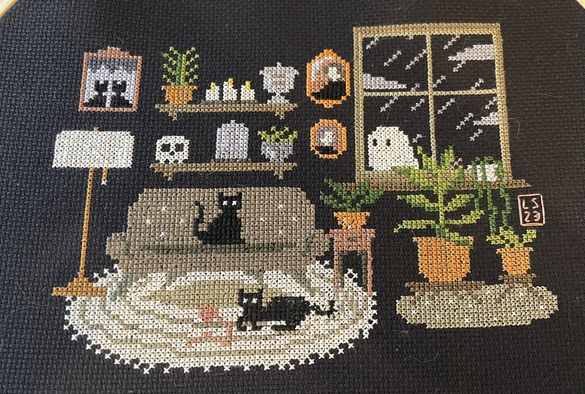 A completed cross-stitch on black Aida of a mid century living room with two black cats lounging and a window where it is raining and there is a ghost looking into the room. From Creepy Cross-stitch.