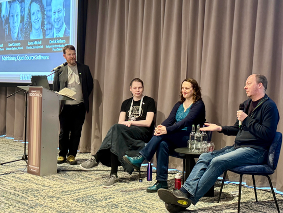 A man (Christopher Hoult) standing at a podium alongside a slide that shows the title Maintaining Open Source Software. To his left are a seated panel of three people: Sam Clements, Lorna Mitchell, and Derick Rethans. Derick has a handheld microphone and is answering a question as the others look towards him.