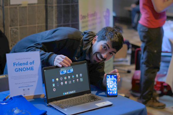 A man is smiling widely, while hunching on a laptop and a phone both running GNOME.

On the desk where the devices stand, there's a page saying "Friend of GNOME" and a pile of GNOME t-shirts.
