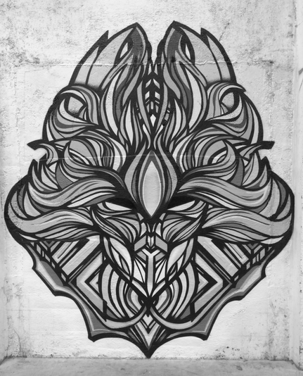 Greyscale image of a vertically symmetrical and stylized mask or demon spirit head painted with acrylic aerosol paint on a rough wall at an indoor bike park. Flowing organic shapes form the hair and face resembling fire with more mechanical forms lying underneath. An almond shaped third eye is the prominent feature above and between the inwardly turned eyes. The image floats on the wall with an empty background.