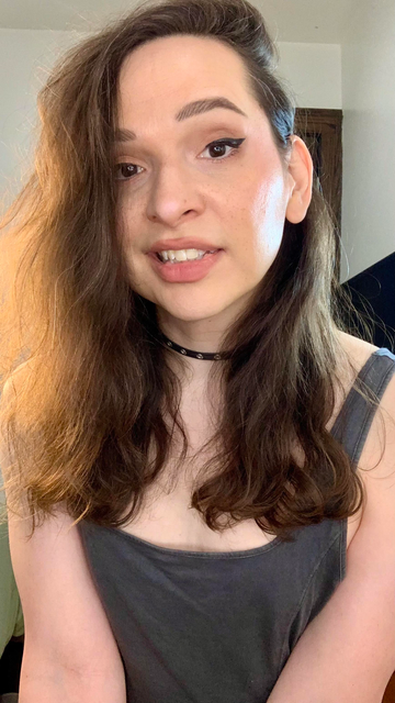 A photo of a cute trans woman (Bijou), smiling, and making eye contact. She has long brown hair, and is wearing lipstick, winged eyeliner, necklace, and a tank top. The photo is set in a room, with warm lighting.