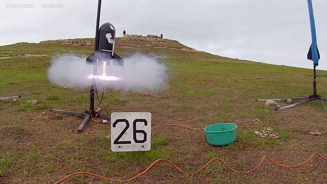 Photo of a stubby, black rocket that looks like Bullet Billy from Nintendo launching from a rail with a jet of high temperature exhaust out the tail end. In the background there are two people watching the launch from a ridge overlooking the rail.