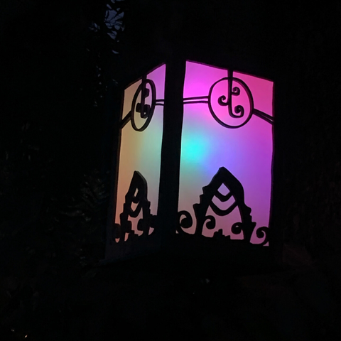 Photo of a box lantern at night, rainbow colors through a woodcut design, leaf & spiral-moon motif