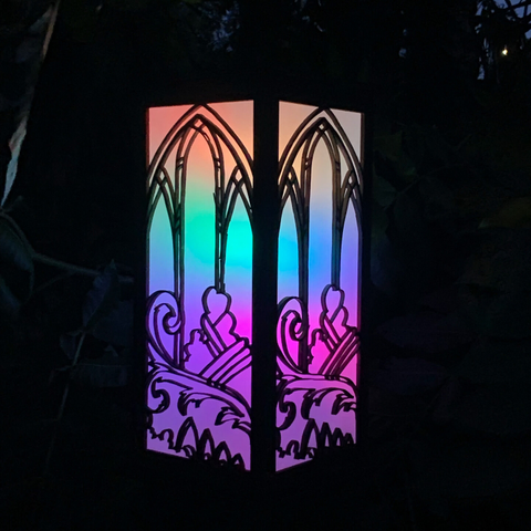 Box lantern at night, gentle rainbow colors through a delicate design of arched windows, plants and ornament