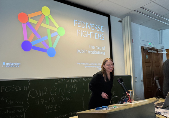 Melanie in an auditorium in front of a microphone and a desk, a slide with the title of the talk and the fediverse sign in the background.