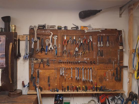 The tool wall of the bike kitchen. Various tools hang on a wooden shelf on the wall. Near the ceiling is bike jousting padded spear.