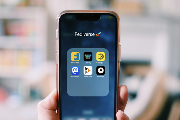 a stock photo for the Fediverse showing a hand that holds a smartphone, where you can see a folder and icons for Friendica, Lemmy, Loops, Mastodon, PeerTube & Pixelfed