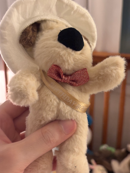 A close-up side view of the same plush Gromit, showing a gold “Happy Birthday” sash across its chest and the white celebratory hat with gold accents.