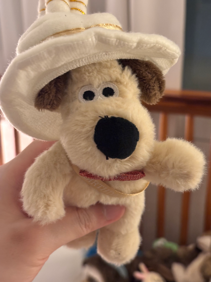 A cream-colored plush Gromit wearing a white birthday hat with gold trim and a pink bow tie. The stuffed toy is being held up against a light background.