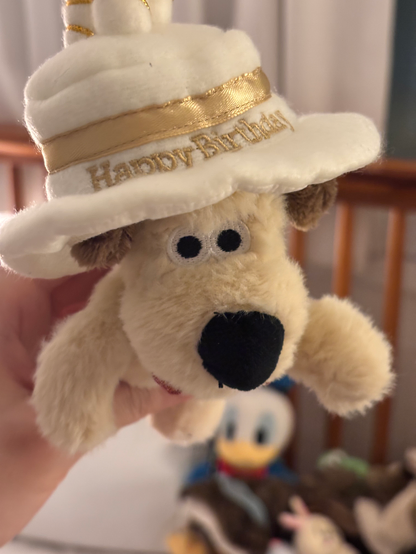 A front-facing close-up of the plush Gromit wearing the white birthday hat with “Happy Birthday” written in gold text. The toy's characteristic black nose and round eyes are clearly visible.