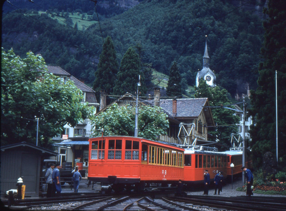 A bright red liveried electric train stands at the entry to a station yard with multiple tracks fitted with racks fanning out.  Behind the train are Swiss chalet style buildings and one ornate clock tower. The background is filled with verdant mountainside.