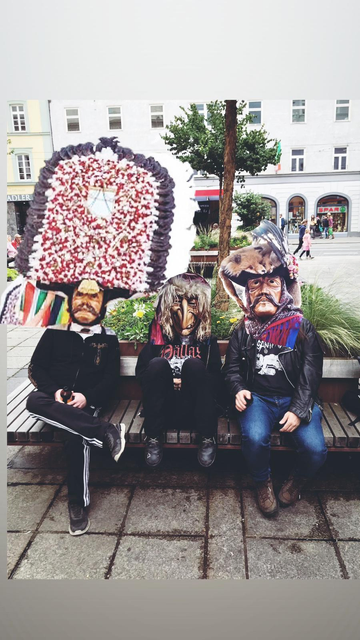 Drei Typen hocken auf einer Bank und mit schlechten Photoshop wurde ihnen Masken aus der Tiroler Fastnacht raufgesetzt. Es sind Holzmasken, die erste zeigt ein Männergesicht samt Schnurrbart und einem riesen Blumenschmuck am Kopf, die zweite eine Hexe, die dritte wieder eine Männermaske mit Schnurbart, aber mit Fuschfell und Federn auf dem Hut