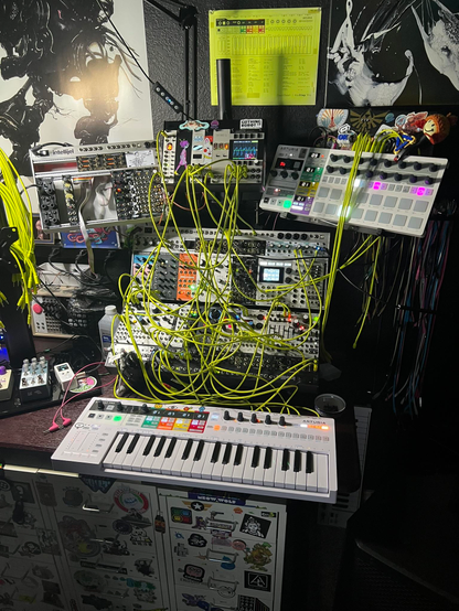 a eurorack synthesizer "tree" - four synths and two keyboards connected via yellow cabling. it looks super messy and tangled, and there a lot of blinky lights and screens.

there are a few guitar pedals nearby too, with ground control's uwu on and a little cat on the screen.