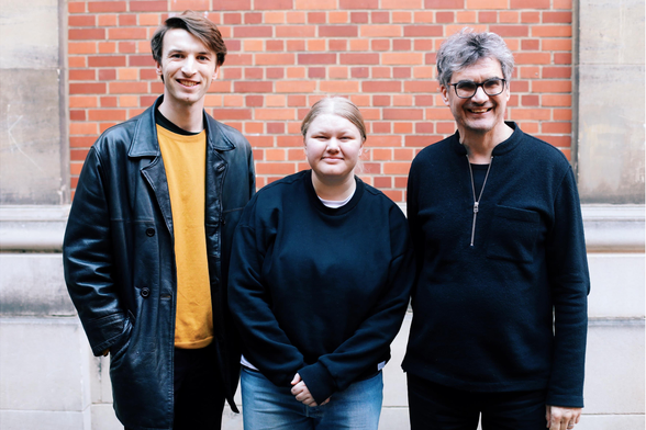 The Newsmast Foundation team. Freddie, in a dark leather jacket and gold jumper. Saskia, in a black jumper and blue jeans. Michael, in black rimmed glasses and a black quarter-zip jumper. 