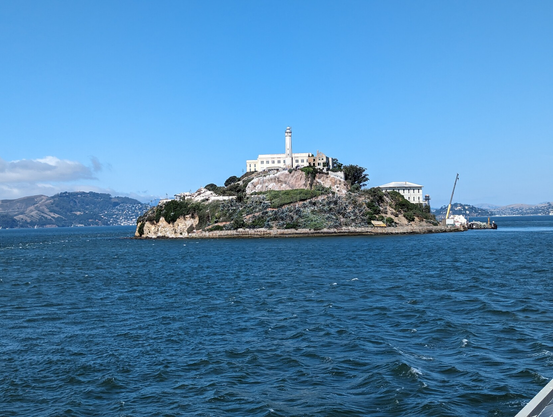 View from the boat to Alcatraz Island looking straight at the lighthouse with the cell block behind it. All in the clear of day with a nearly cloudless blue sky behind it.